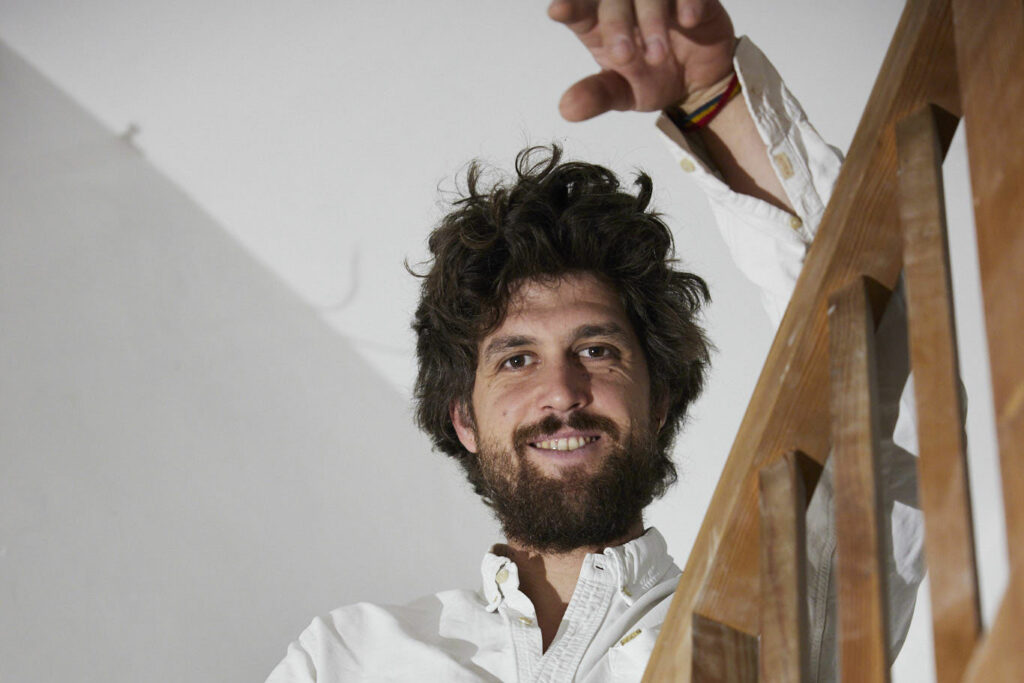 Das Foto zeigt ein Portrait des Künstlers Alexander Wierer. Er lächelt in die Kamera, während er seinen linken Unterarm auf einem Treppengeländer abstützt.