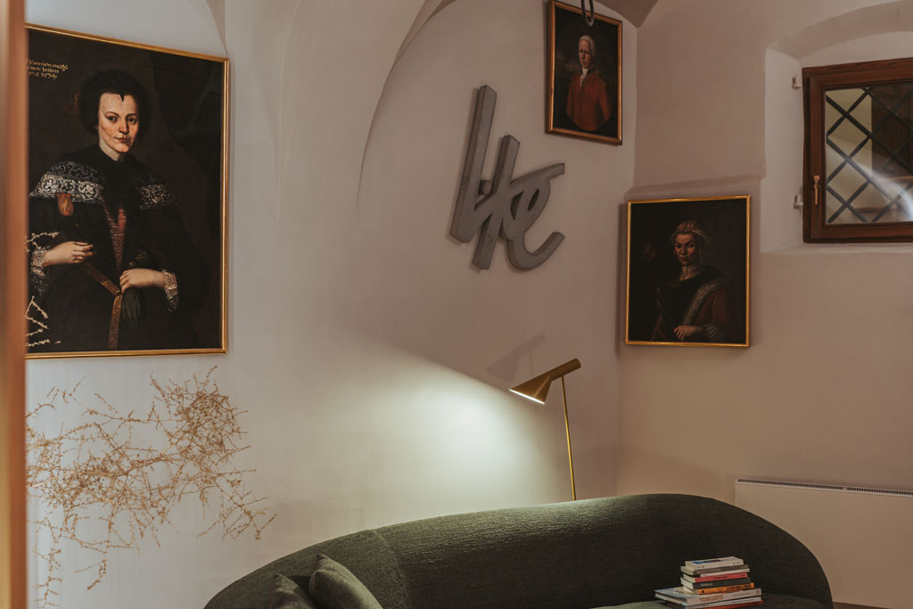 Das Foto zeigt einen Bereich im Erdgeschoss des Lasserhaus. In der Ecke des Raumes steht ein grünes Sofa. An der Wand hängen drei klassische Gemälde aus der Kunstsammlung des Lasserhaus, dazwischen hängt das Werk "like" von Petra Polli.