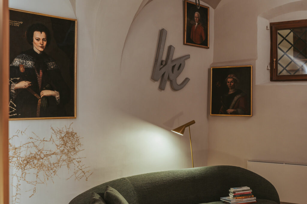 Das Foto zeigt einen Bereich im Erdgeschoss des Lasserhaus. In der Ecke des Raumes steht ein grünes Sofa. An der Wand hängen drei klassische Gemälde aus der Kunstsammlung des Lasserhaus, dazwischen hängt das Werk "like" von Petra Polli.