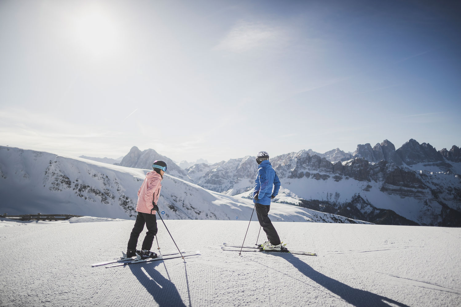 Zwei Personen in Winterkleidung stehen auf einem verschneiten Berghang mit Skiern und genießen das Skifahren auf der Plose bei Brixen bei strahlendem Sonnenschein, mit weit entfernten schneebedeckten Gipfeln und einem klaren blauen Himmel im Hintergrund.