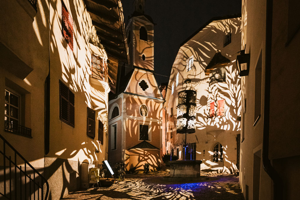 Eine schmale Kopfsteinpflasterstraße wird nachts während des Brixen Waterlight Festivals von komplizierten Lichtprojektionen mit Blattmustern beleuchtet, die dramatische Schatten auf die Gebäude werfen und einen kleinen zentralen Brunnen umgeben.