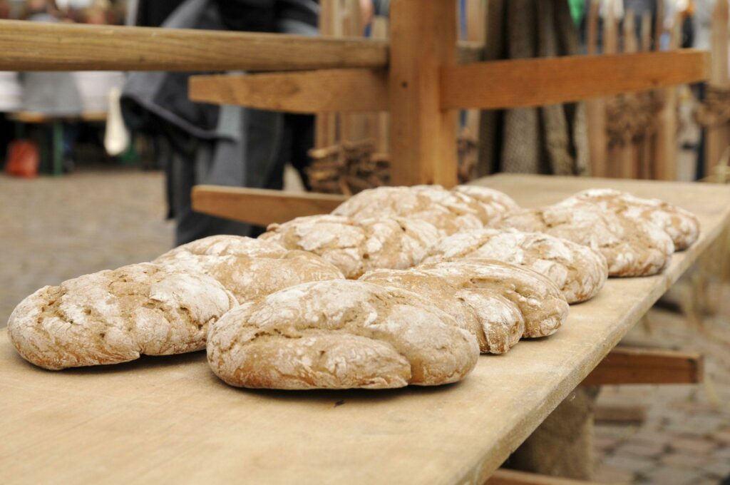 Mehrere runde Brote mit rustikaler Kruste liegen in Reihen auf einem Holztisch im Freien auf dem Brot- und Strudelmarkt am Domplatz Brixen. Im verschwommenen Hintergrund sind weitere Brote und Holzkonstruktionen zu sehen.