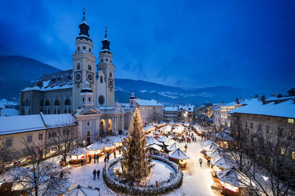 Der verschneite Brixner Domplatz in der Abenddämmerung mit einem großen Weihnachtsbaum in der Mitte, umgeben von festlichen Marktständen und Menschen. Der Weihnachtsmarkt Brixen in Südtirol erstrahlt neben einer großen Kirche mit Zwillingstürmen unter dem blauen Abendhimmel.