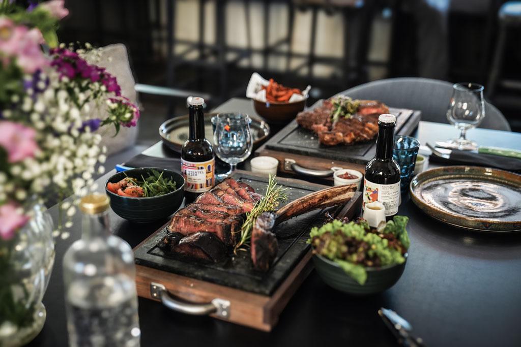 Das Bild zeigt einen gedeckten Tisch im Restaurant Alter Schlachthof in Brixen. Auf dem Tisch befinden sich ein Steak, Salate, das hauseigene Viertel Bier und Süsskartoffel Spalten.
