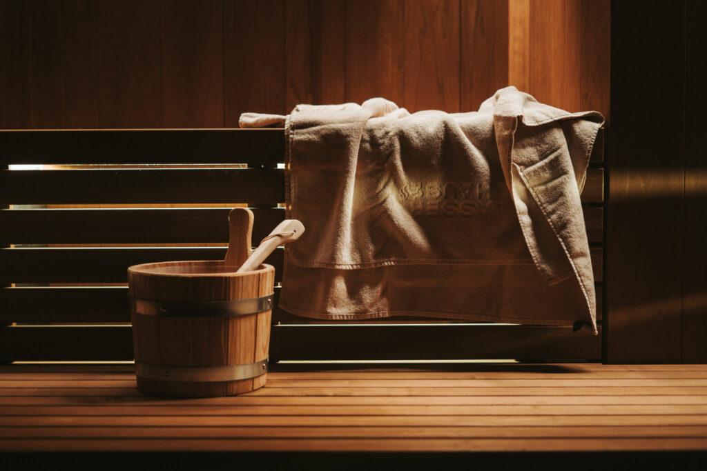Ein Handtuch, ein Aufgusskübel und eine Schöpfkelle in der Sauna des Arthotel Lasserhaus in Brixen bei wohlig warmer Lichtstimmung.