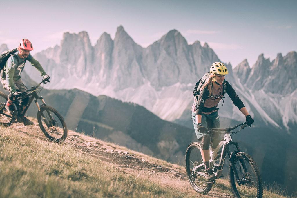 Zwei Radfahrer fahren mit ihren Mountainbikes auf einem unbefestigten Weg in der Nähe der Plose Brixen, mit dramatischen, zerklüfteten Bergen und einem klaren Himmel im Hintergrund. Beide tragen Helme und Rucksäcke und genießen ein aufregendes Outdoor-Abenteuer.